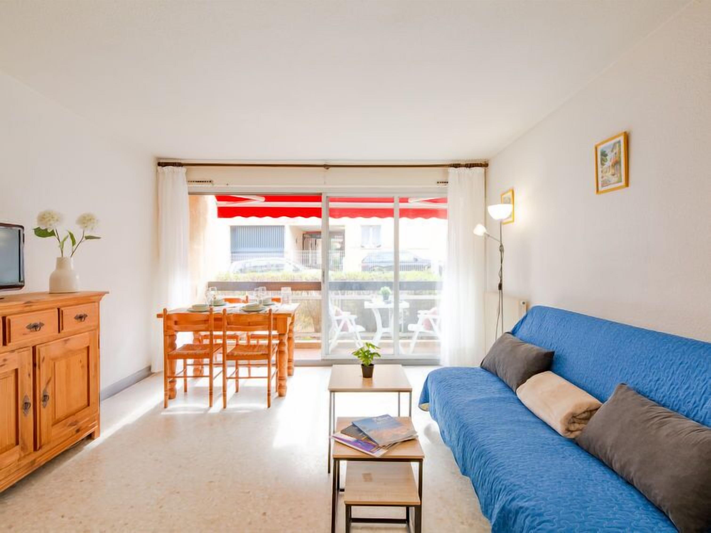Ein Zimmer mit blauem Sofa, Esstisch aus Holz und Stühlen, einem kleinen Tisch mit Pflanze und Büchern sowie einem Fernseher auf einer Holzkommode.