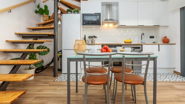 A modern kitchen with white cabinets, a wooden staircase, and a dining area with metal chairs.