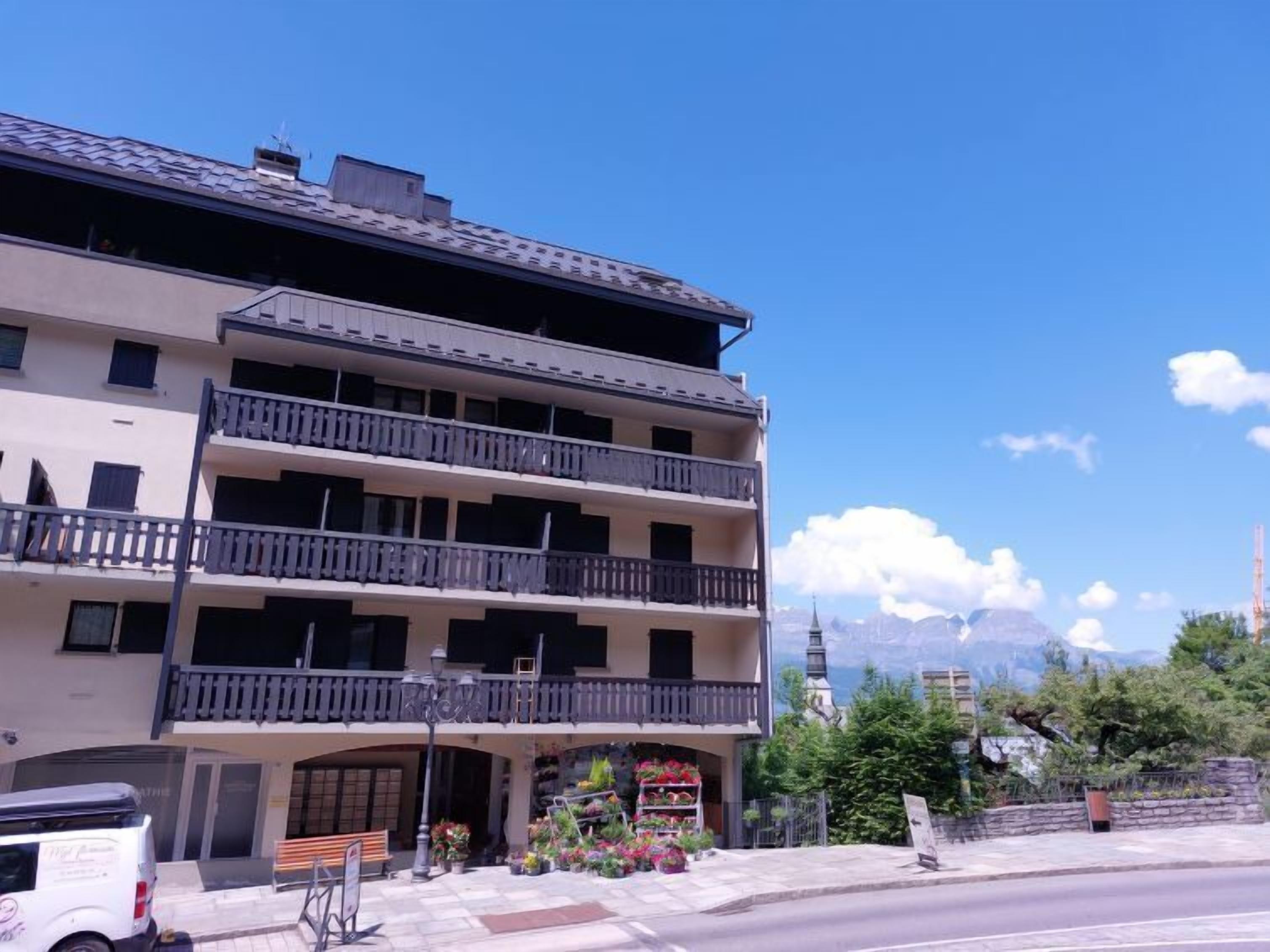A multi-story building with balconies, a flower shop on the ground floor, and a clear view of mountains in the background.