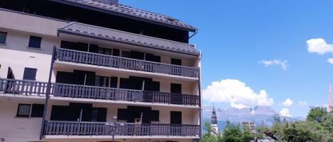 A multi-story building with balconies, a flower shop on the ground floor, and a clear view of mountains in the background.