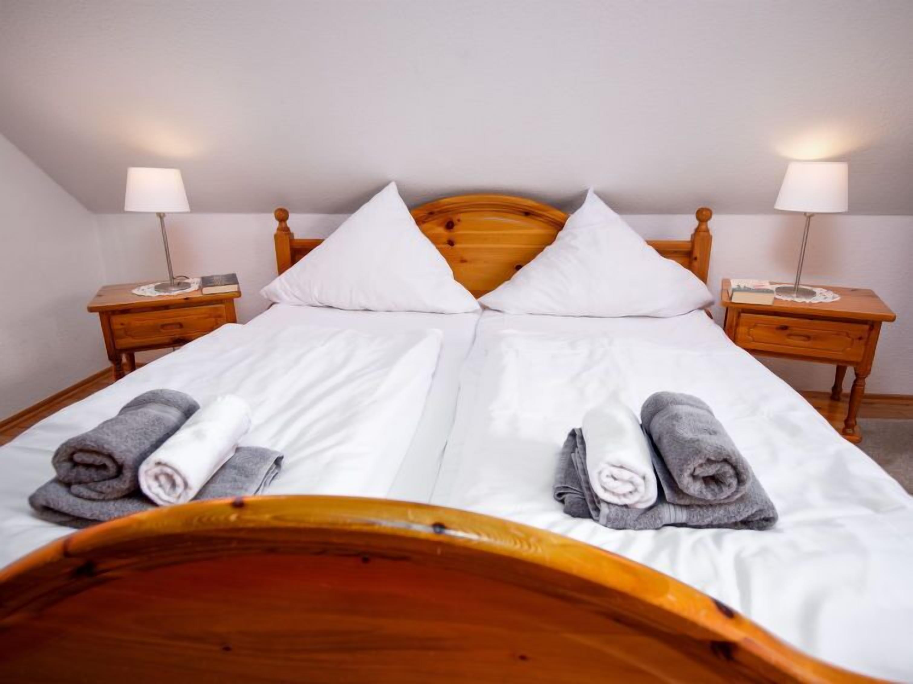 Ein ordentlich bezogenes Bett mit weißen Leinen und zwei Kissen, an beiden Seiten mit Holznachtischen, auf denen Lampen und Bücher stehen.