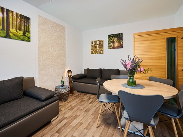 Ein modernes Wohnzimmer mit einer Couch, einem Couchtisch und einem Esstisch mit Stühlen. An den Wänden sind Dekorationen und ein Holztür.