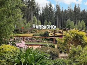 A sign reading "HARRACHOV" in a forested area with a wooden bridge and various plants. - Helena Harrachov by Interhome (Harrachov)