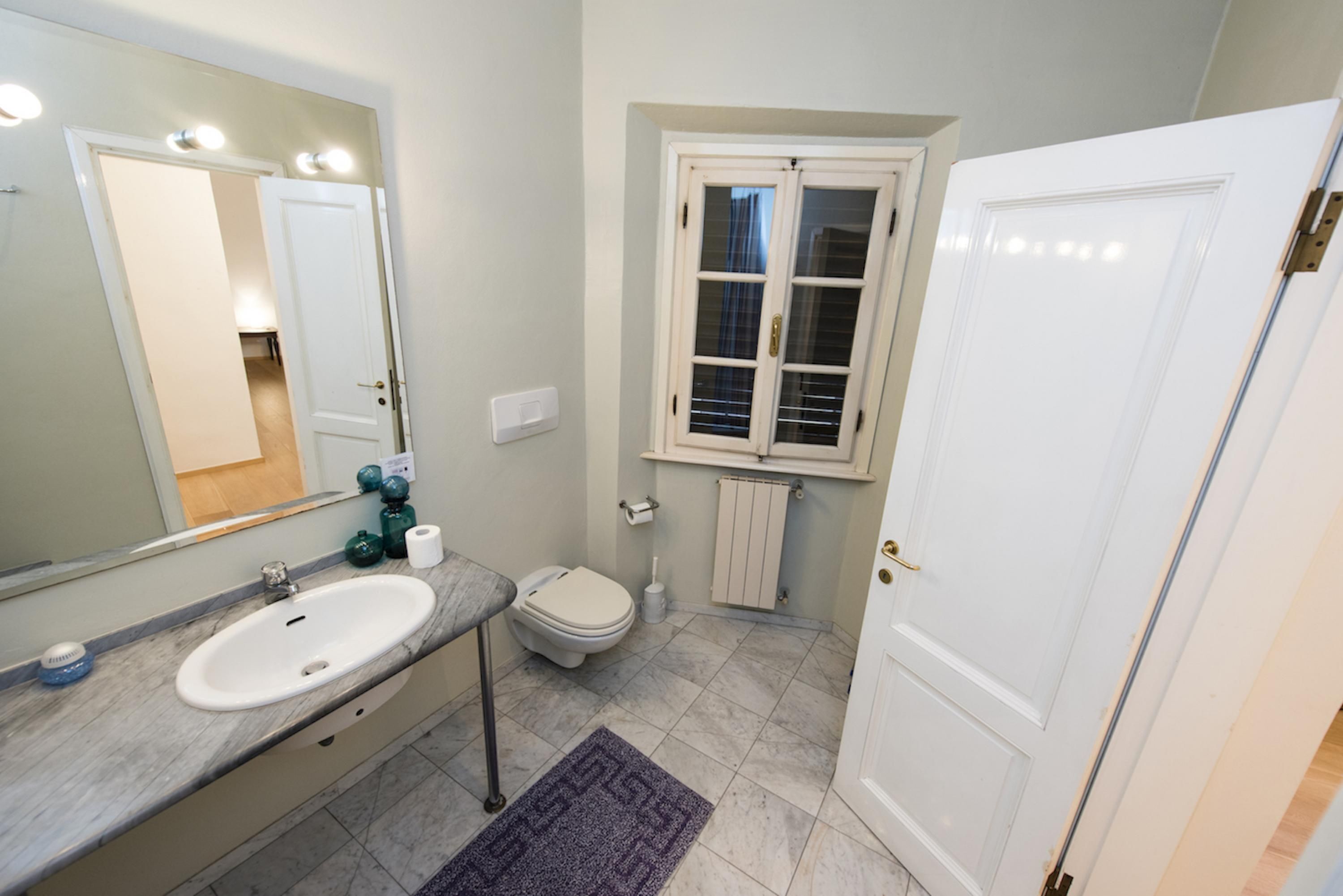 Un baño con lavamanos blanco, espejo, inodoro y una ventana con persianas.