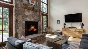 A spacious living room with a stone fireplace, comfortable seating, a flat-screen TV, and a large window offering a view of the outdoors. - Month long Rental- Teton Springs Home 4 Bedroom (Victor)