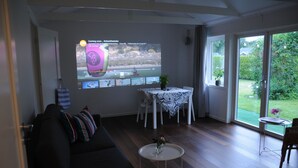A living room with a large screen displaying a video, a black sofa with striped cushions, a small round table with a vase, and a view of a garden through glass doors.