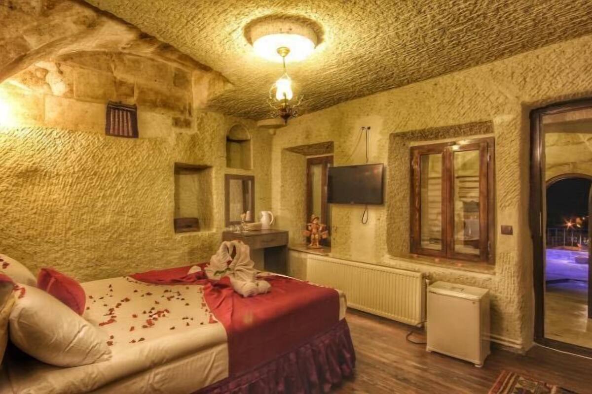 Una habitación con pared de piedra, cama con ropa de cama roja, una mesita y un televisor.