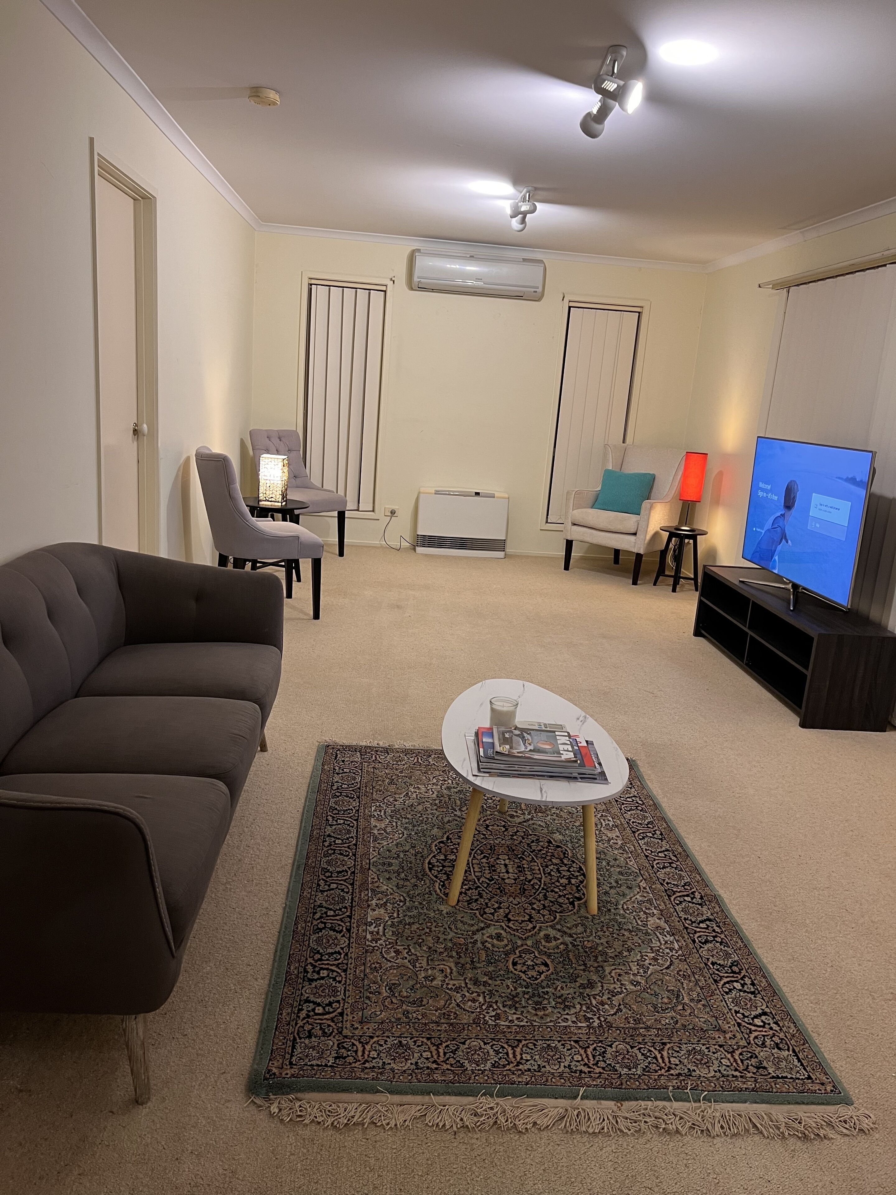 A living room with a dark brown sofa, a white coffee table with magazines, a patterned rug, a TV, and a ceiling fan.