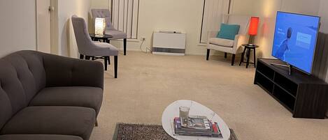 A living room with a dark brown sofa, a white coffee table with magazines, a patterned rug, a TV, and a ceiling fan.