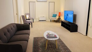 Una sala de estar con un sofá marrón, una mesita blanca con revistas, un televisor sobre un mueble bajo y una alfombra estampada.