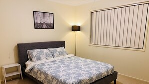 Un dormitorio con una cama, una mesita de noche, una lámpara de pared, una ventana con persianas y un cuadro en la pared.