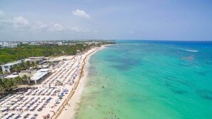 Vista aerea di una spiaggia con file di ombrelloni e lettini, un mare cristallino e una costa lontana con edifici.