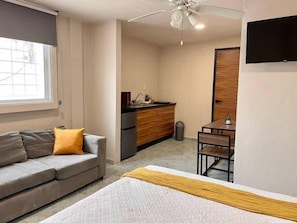 A bedroom with a bed, a gray sofa with a yellow pillow, a small table, and a kitchenette area with a sink and cabinets. - Cozy Studio | Downtown Cancun | WiFi & AC (CancÃºn)
