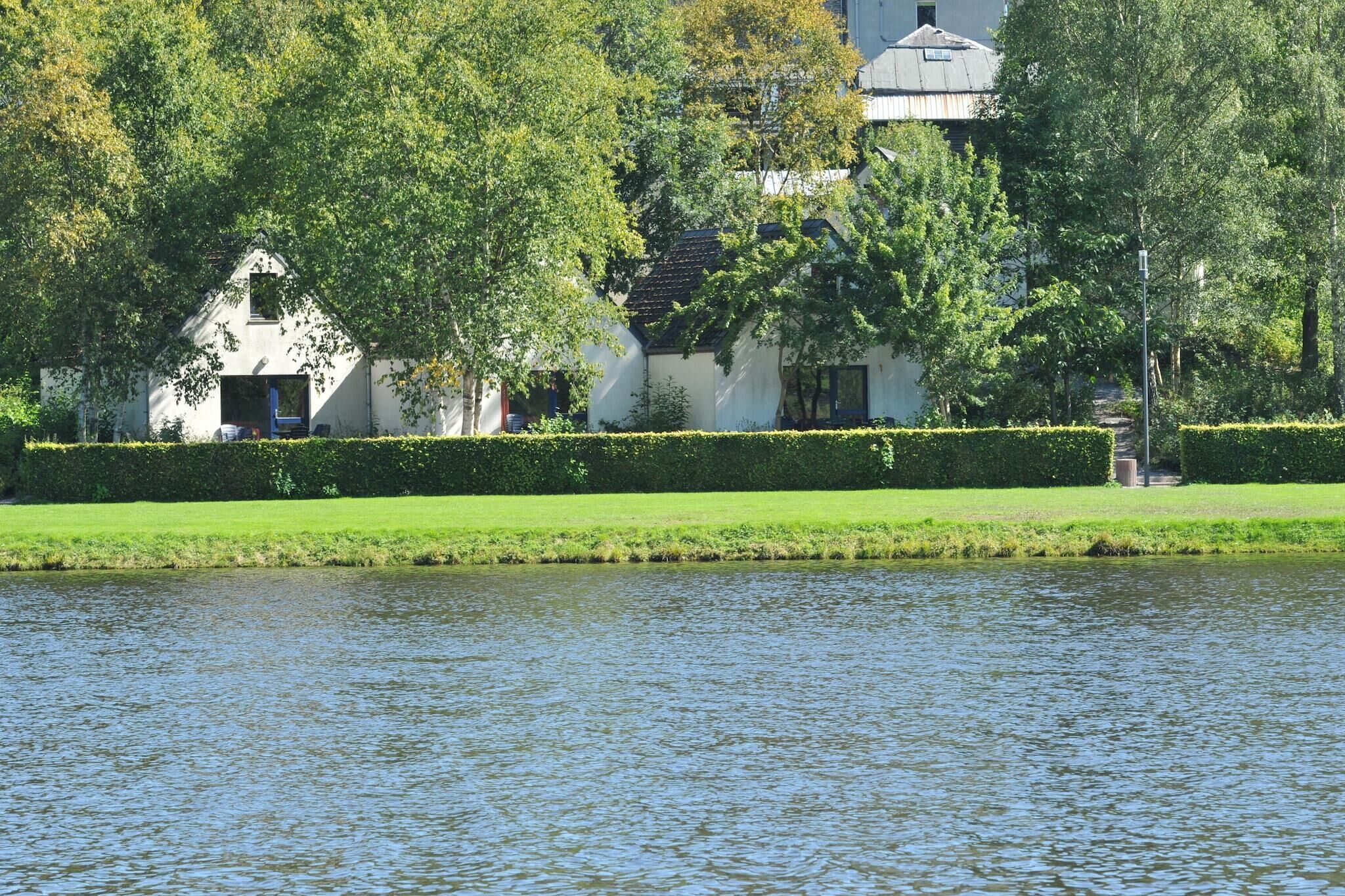 Une vue paisible sur la rivière, avec une maison blanche, des haies soigneusement taillées et une végétation luxuriante.