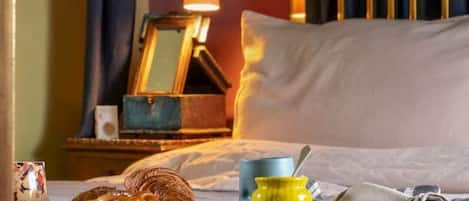 Ein ordentlich bezogenes Bett mit Decke, eine Nachttischlampe, ein kleiner Tisch mit Tasse und Glas sowie ein Fenster mit Vorhängen.