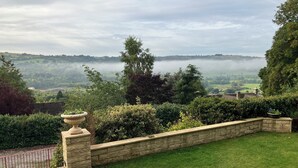 Un jardín ajardinado con un muro de piedra, un césped y vistas de un valle cubierto de niebla sobre las colinas.