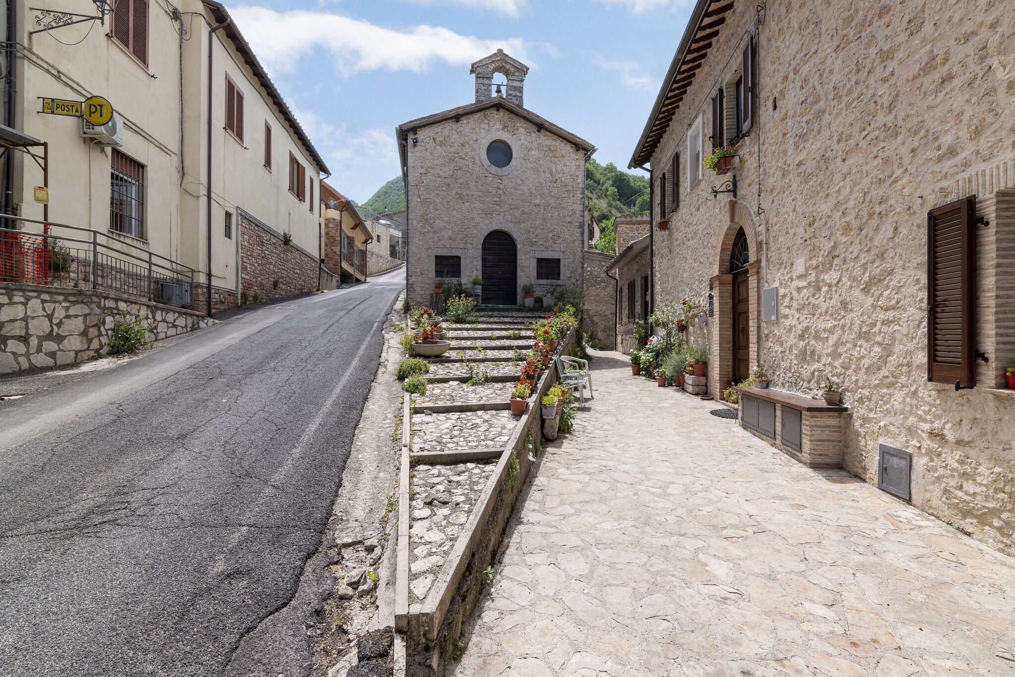 A narrow street with stone buildings, a church with a bell tower, and a paved sidewalk with flower beds.