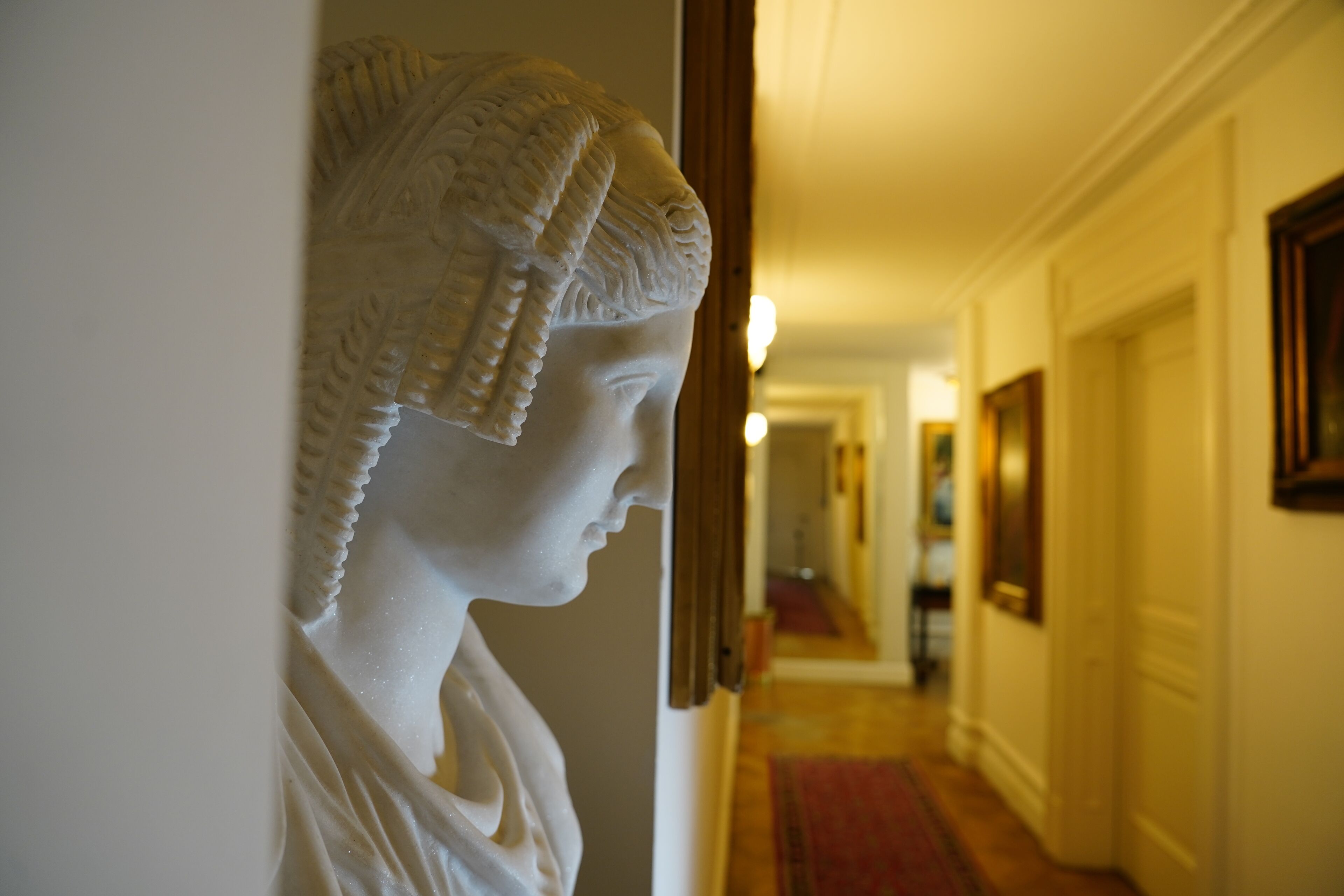 A hallway with a classical sculpture on the wall, a red carpet, and framed artwork on the walls.