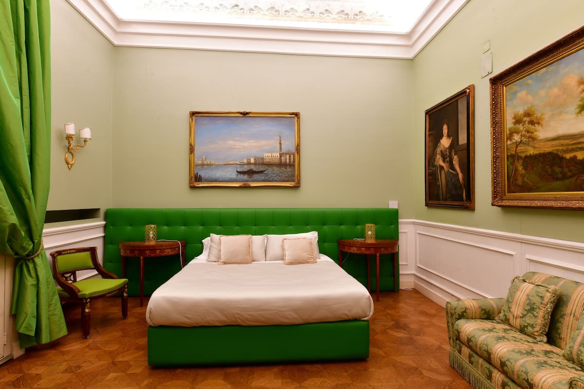 A room with a green upholstered bed, a green armchair, a wooden chair, and two small tables with lamps.