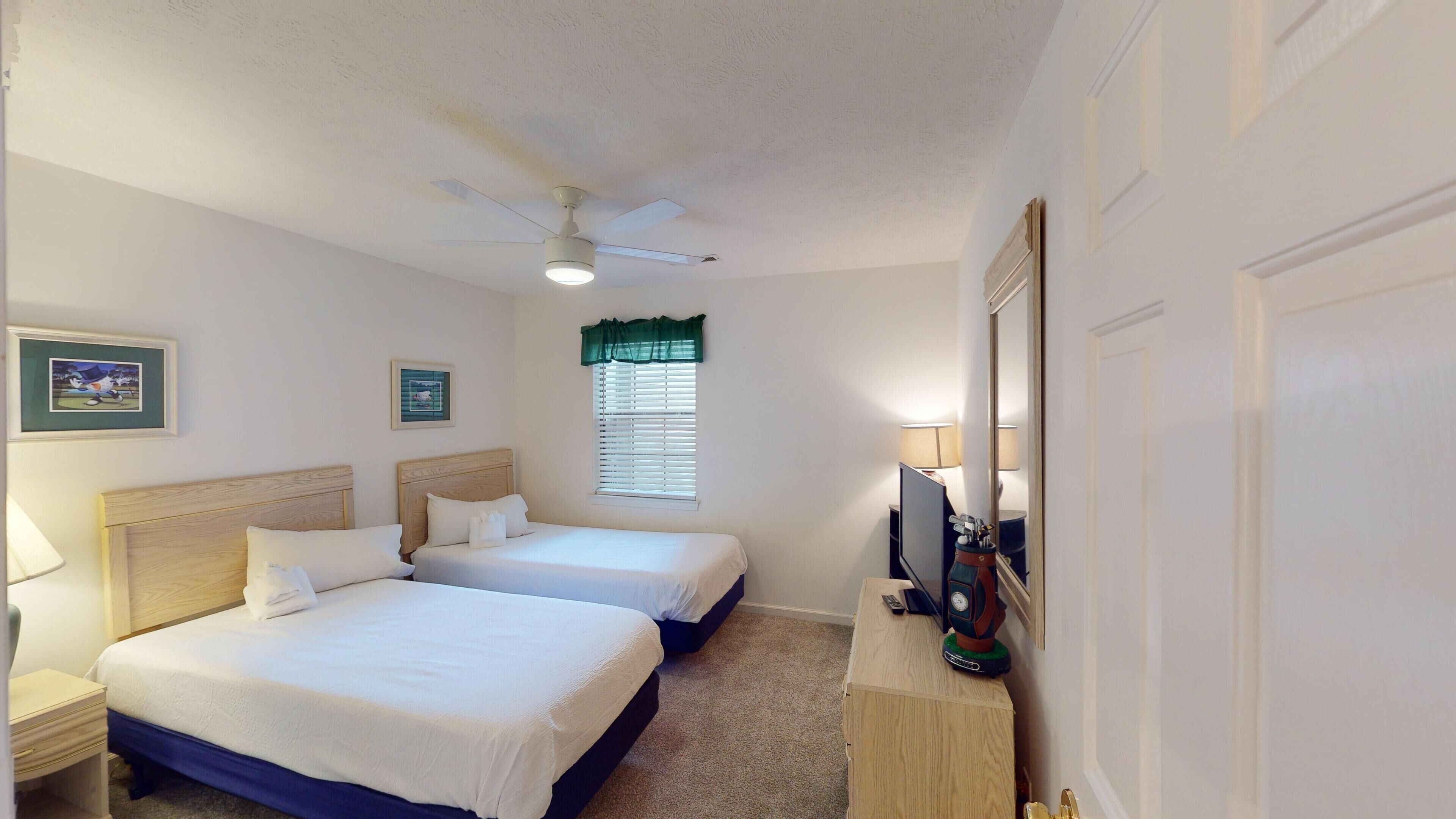 A hotel room with two beds, a ceiling fan, a window with blinds, a TV on a stand, and a door.