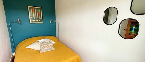 Un dormitorio pequeño con una cama, pared azul, un cuadro enmarco y un espejo.