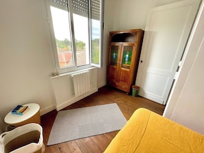 A room with a radiator, a cabinet, a window, a small table, a chair, and a yellow bed.