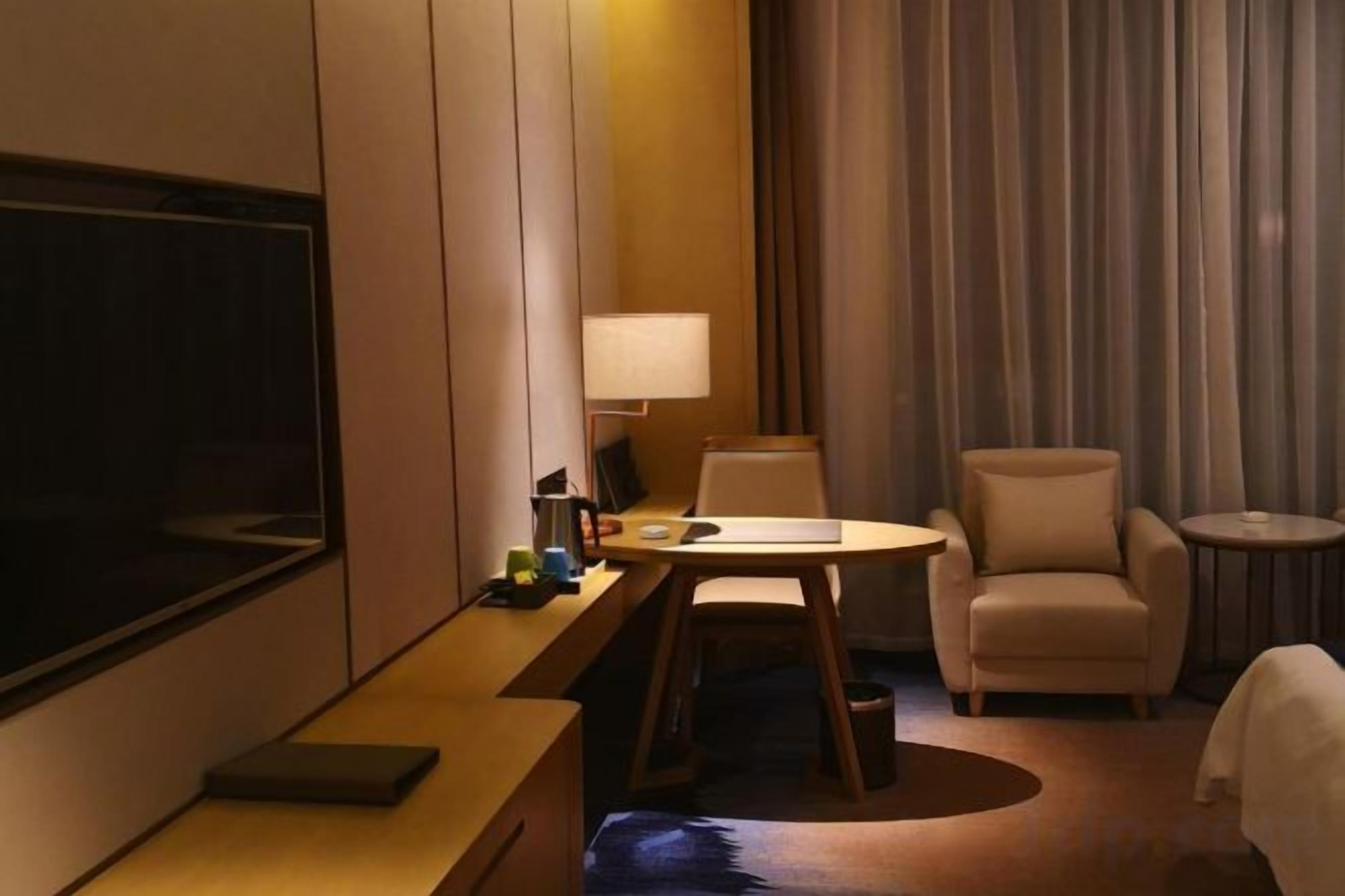 A hotel room with a flat-screen TV, a desk with a lamp, a chair, a round table, and a bed.