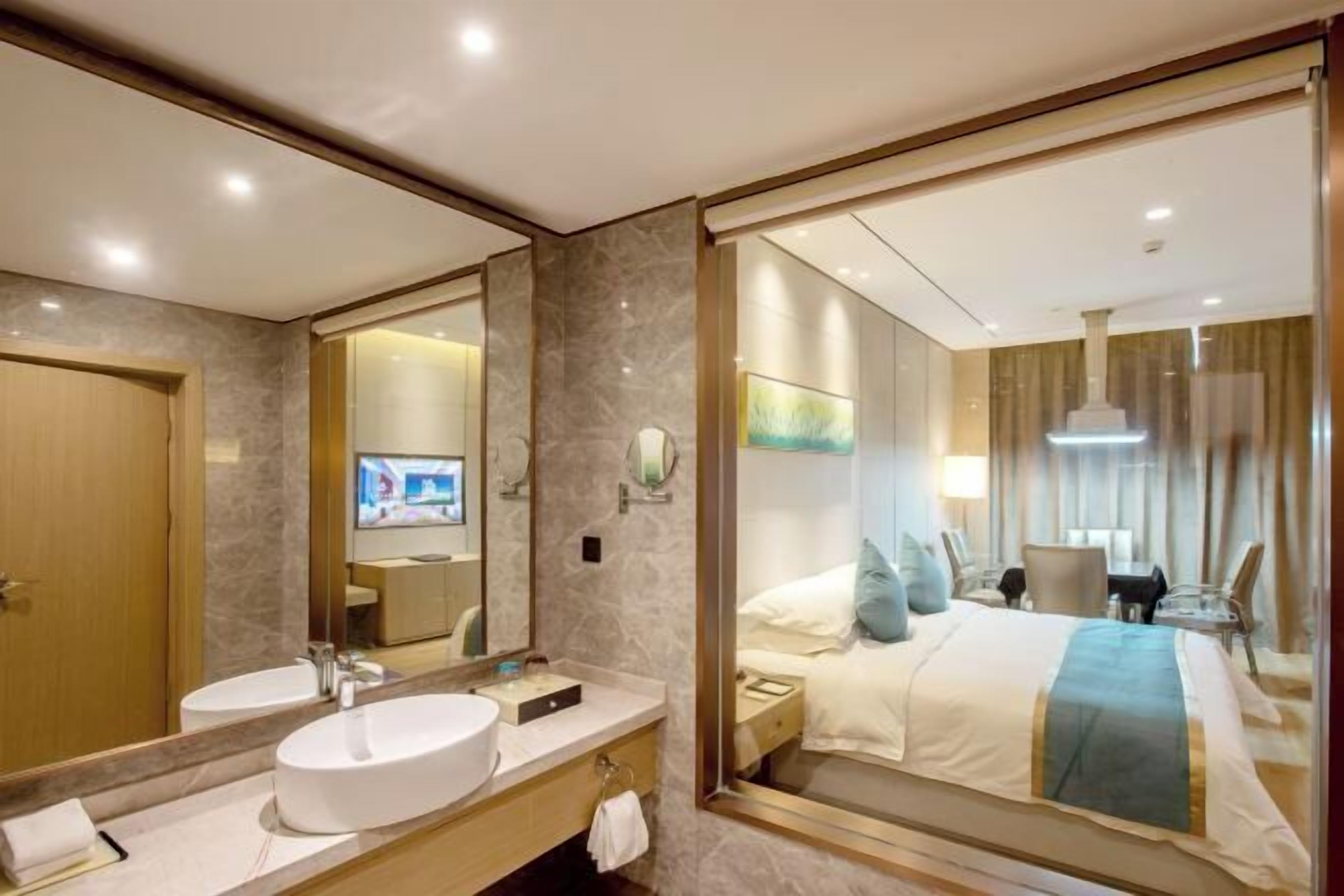 A hotel room with a large mirror, a round white sink, a bed with a blue and white bedspread, and a television in the background.