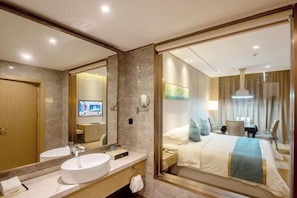A hotel room with a large mirror, a round white sink, a bed with a blue and white bedspread, and a television in the background.