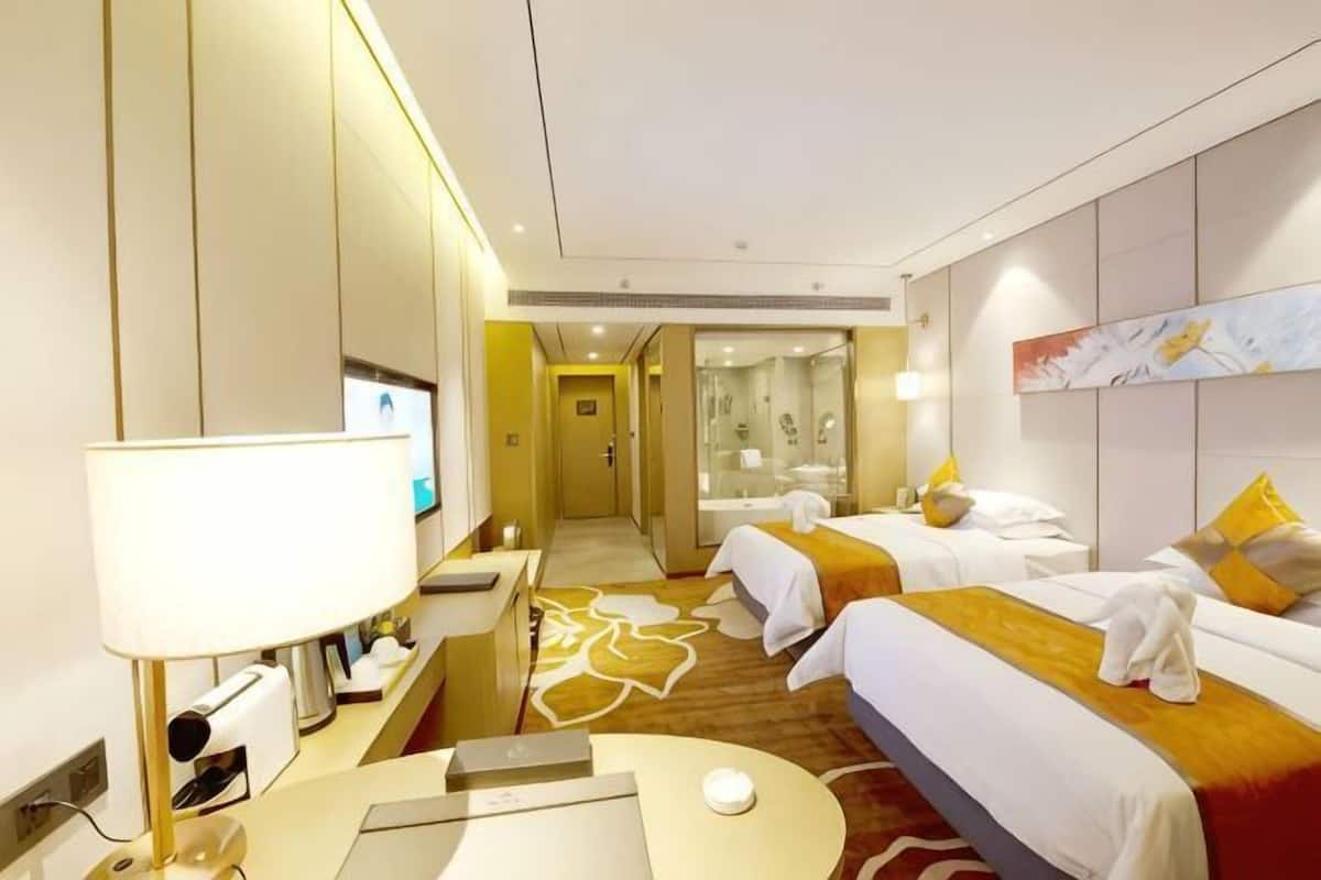 A hotel room with two beds, a desk with a lamp, a TV, and a bathroom with a shower and mirror.