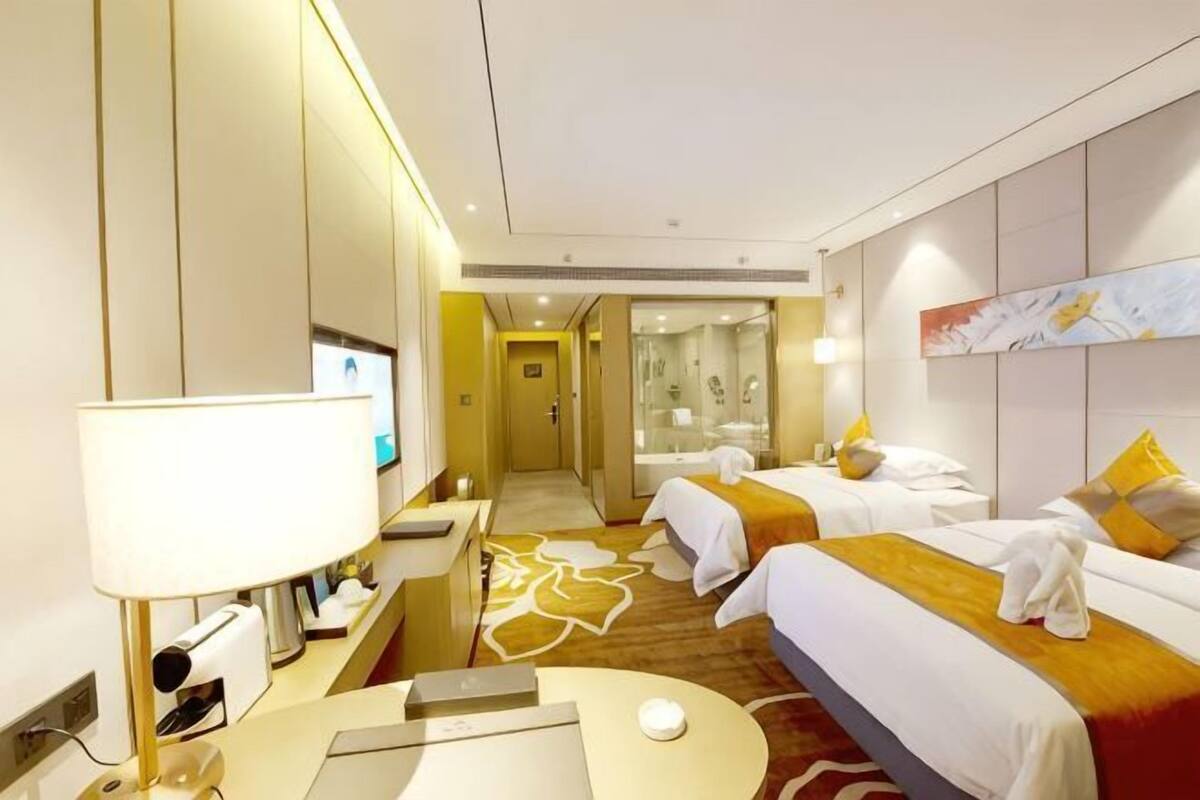 Habitación de hotel con dos camas, un escritorio con lámpara, televisor y baño con ducha y espejo.