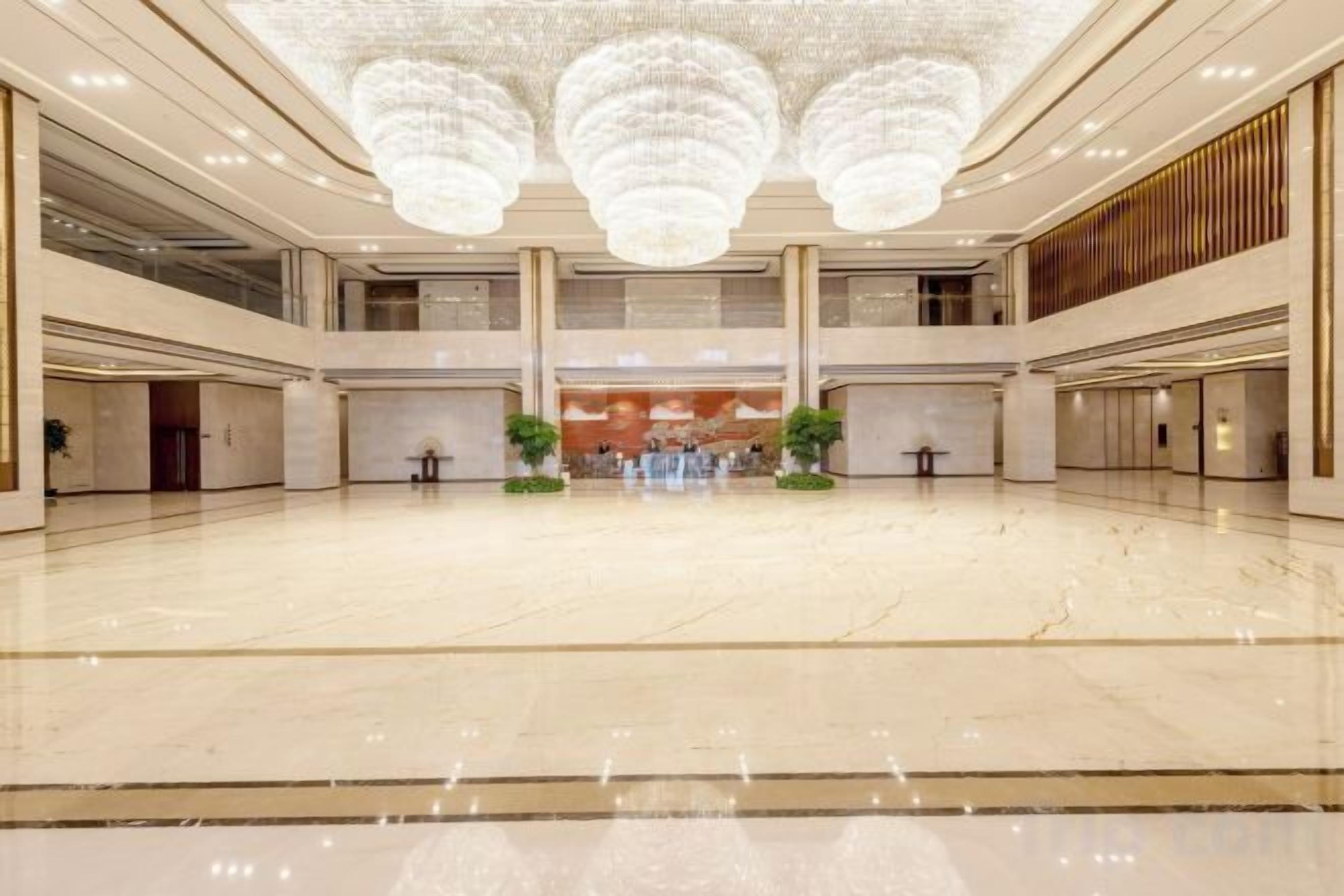 A spacious lobby with a high ceiling and chandeliers, featuring a central reception desk and potted plants.
