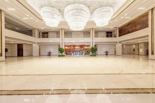 Um hall espaçoso com teto alto e lustres, destacando um balcão de recepção central e plantas em vasos.