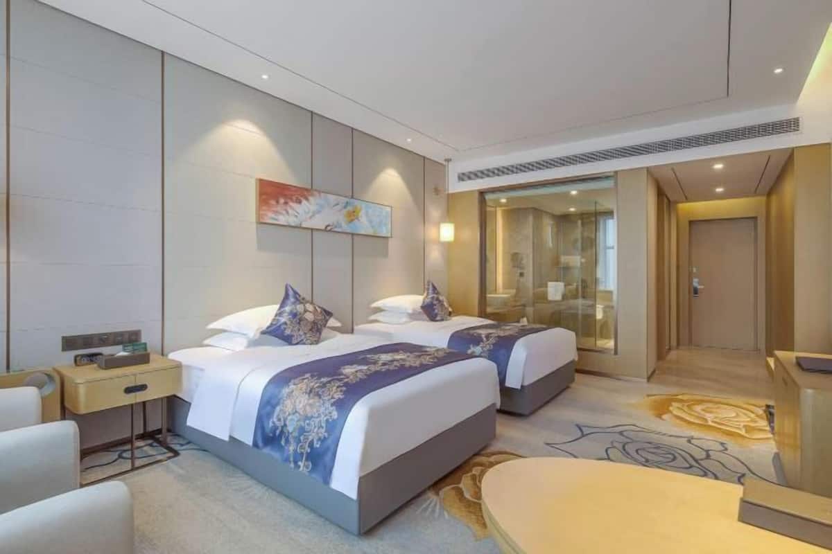 A hotel room with two beds, a desk, a chair, a painting on the wall, and a glass door leading to a bathroom.