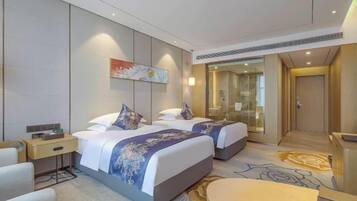 A hotel room with two beds, a desk, a chair, a painting on the wall, and a glass door leading to a bathroom.