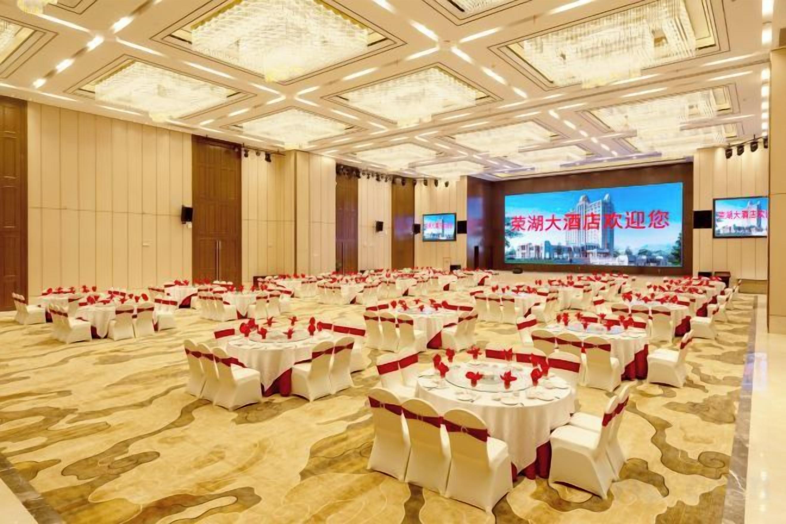 A banquet hall with multiple round tables, each with a white tablecloth and red sashes, arranged in rows.