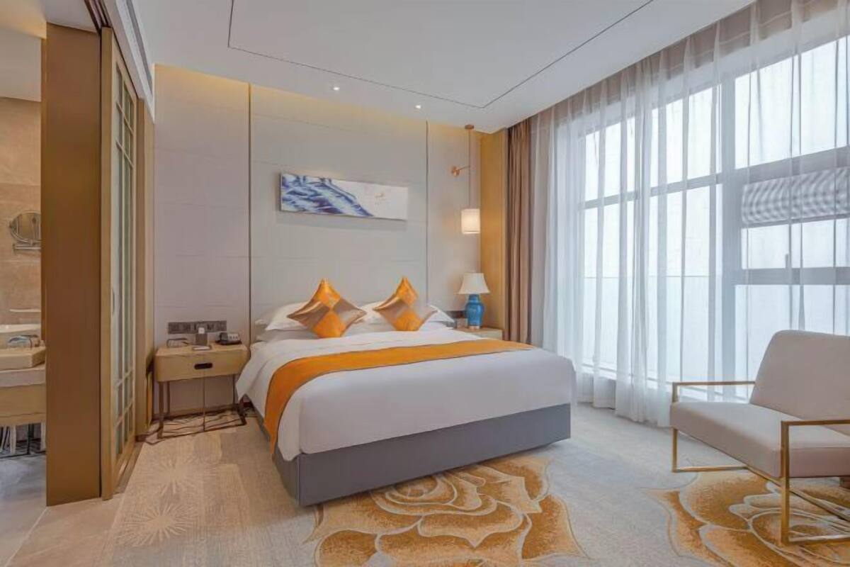 A hotel room with a bed, a nightstand, a chair, a large window with curtains, and a wall-mounted artwork.