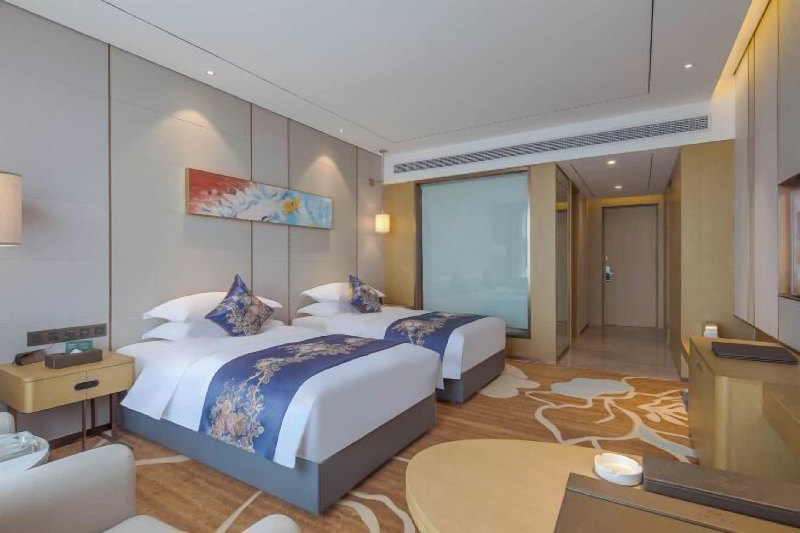 A hotel room with two beds, a desk, a chair, a painting, and a reception area.