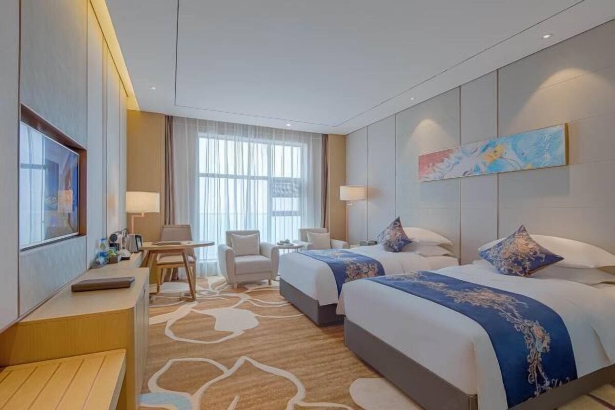 A hotel room with two beds, a desk, a chair, a TV, and a large window with curtains.