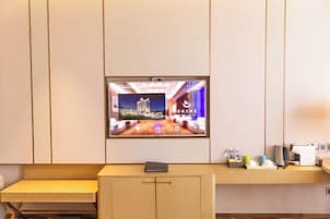 Habitación de hotel con televisión de pantalla montada que muestra un paisaje urbano, una mesita con un libro y una encimera con varios objetos.