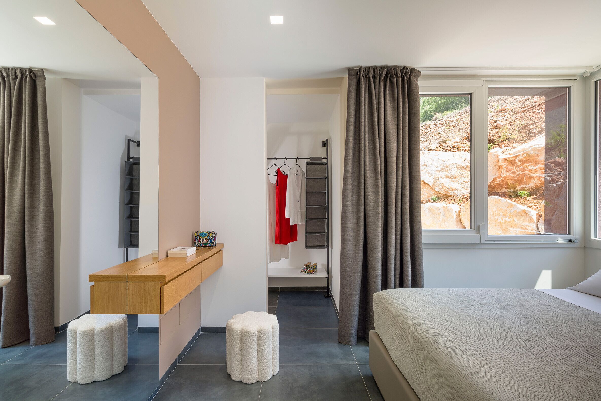 A modern hotel room with a bed, a wooden desk, two ottomans, a wardrobe, and a window with curtains.