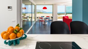 Une cuisine moderne avec un plan de travail en marbre, un saladier rempli d’oranges et une vue sur un coin repas agrémenté d’un luminaire suspendu rouge et d’une table avec des chaises.
