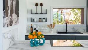 Une cuisine moderne avec un plan de travail en marbre, un saladier rempli d’oranges, une bouteille de vin et une fenêtre donnant sur un paysage verdoyant.