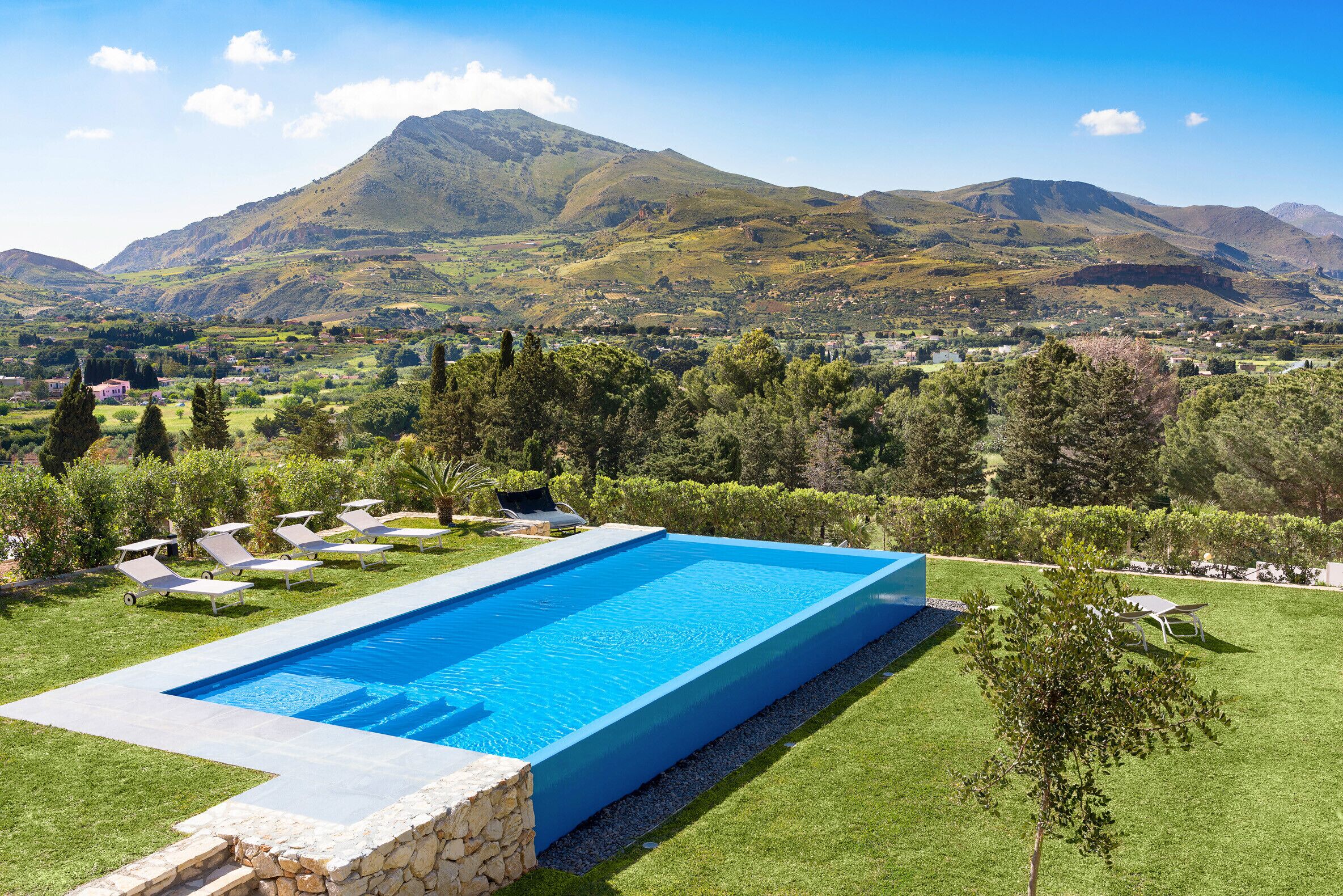 A rectangular swimming pool with a stone border, surrounded by a well-maintained lawn and trees.