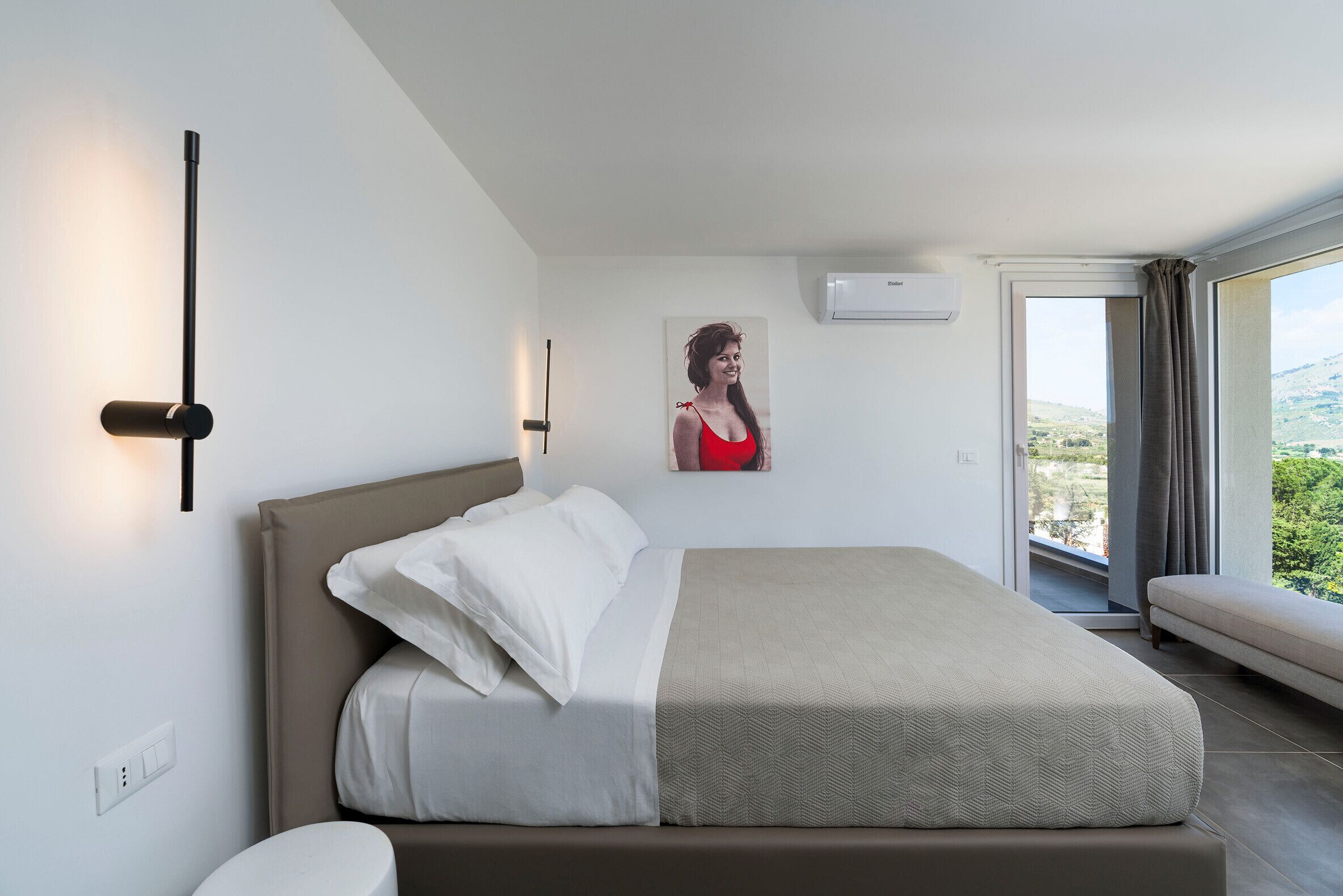 A modern bedroom with a bed, a wall-mounted air conditioner, and a window with a view of greenery.