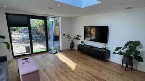 Un salón moderno con tragaluces, suelo de madera, un otoman rosa, un televisor en la pared y un mueble de entretenimiento negro.