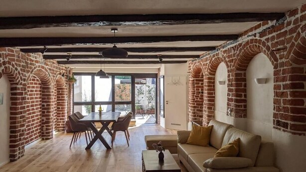 Ein großzügiger Wohnbereich mit Holzboden, einer Couch, einem Couchtisch und einem Esstisch mit Stühlen. Backsteinbögen und ein großes Fenster sind zu sehen.