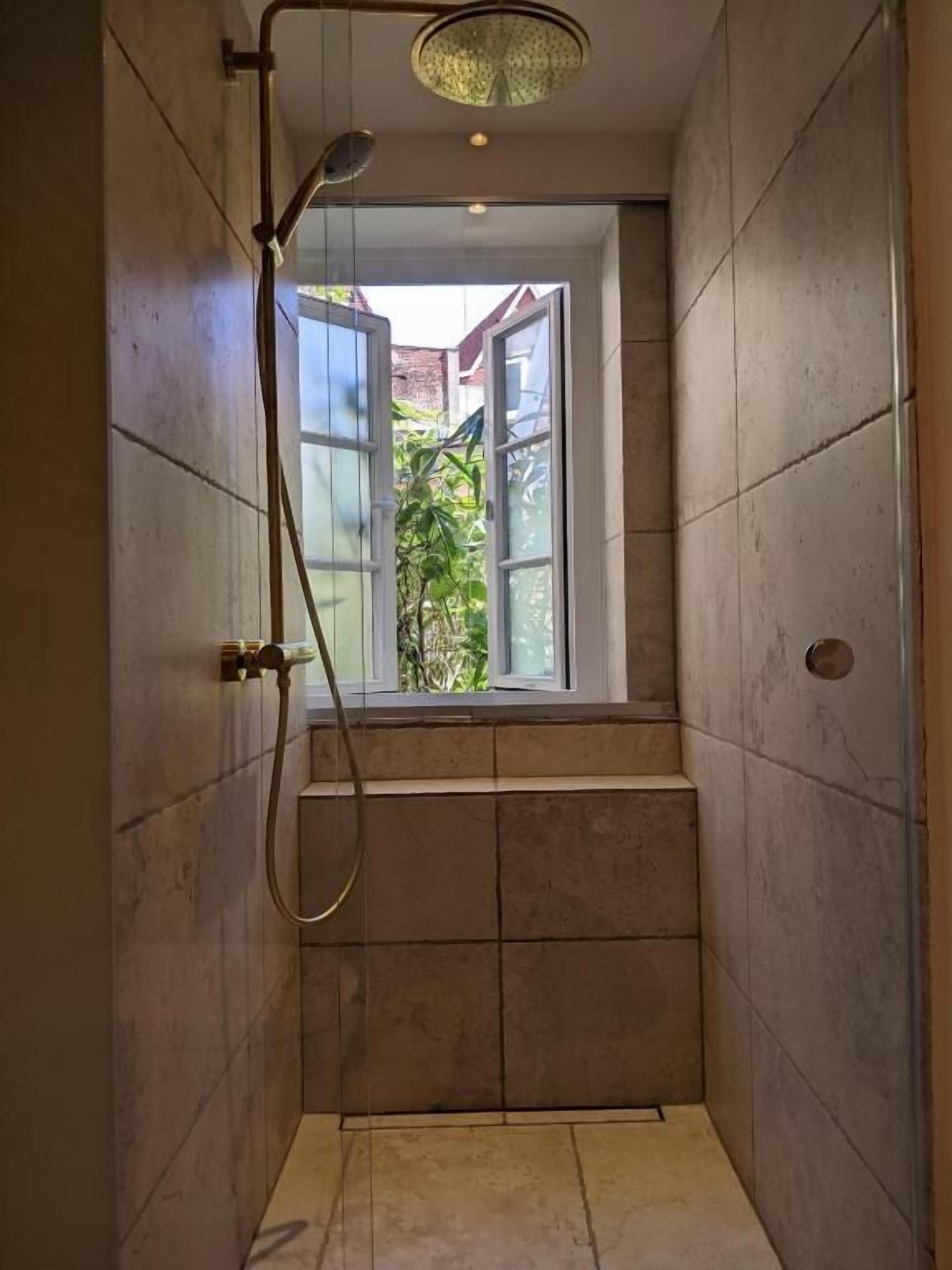 Un petit espace douche fermé, doté d’une fenêtre, de murs carrelés et d’une plante visible à l’extérieur.