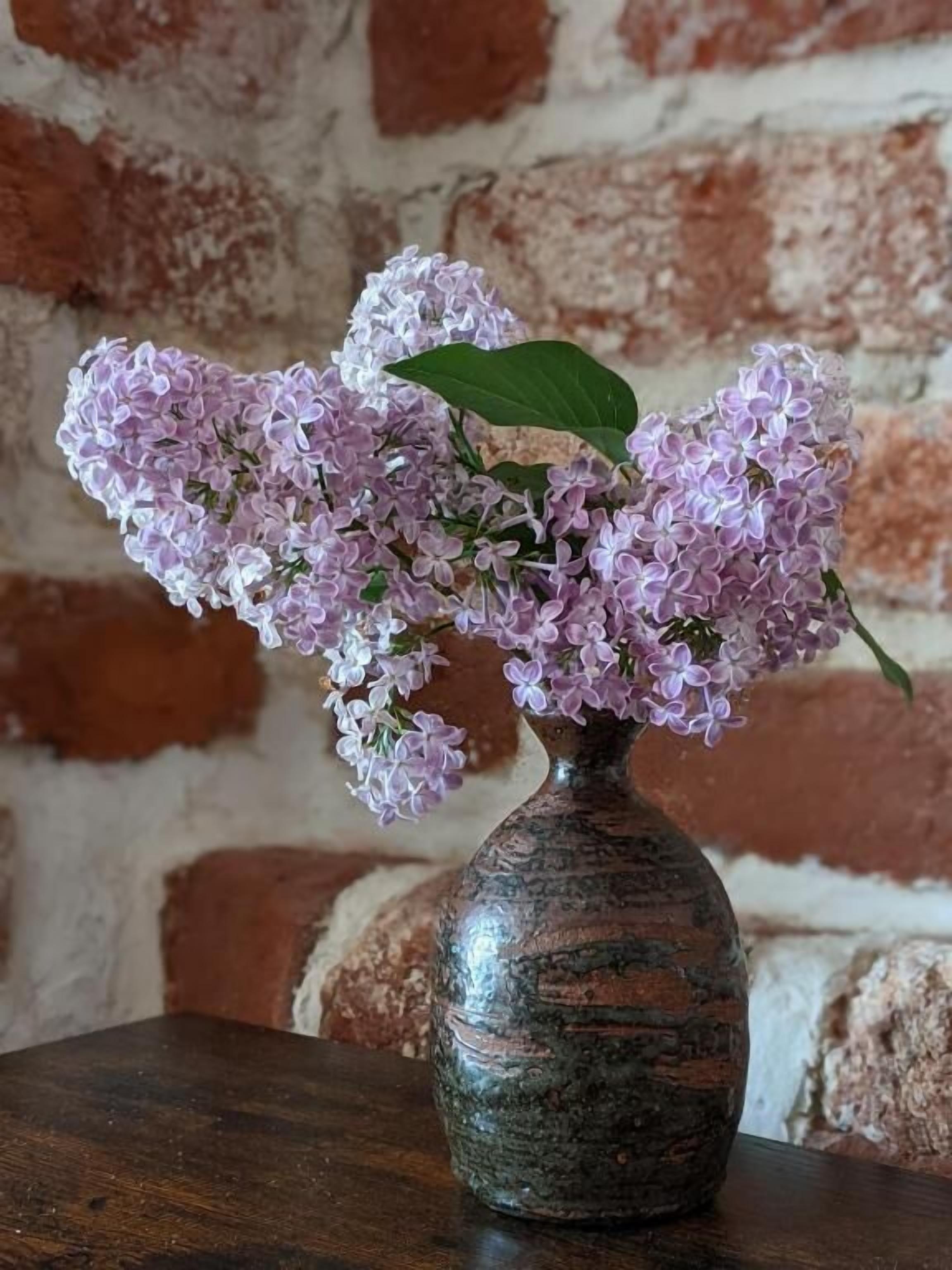Eine Vase mit Fliederblüten auf einem Holztisch vor einer Backsteinwand.