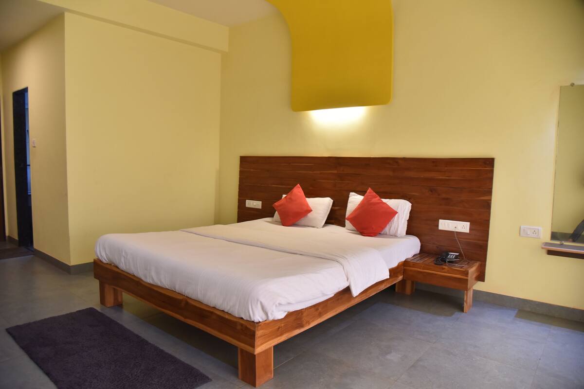 Ein Schlafzimmer mit einem hölzernen Bett, weißer Bettwäsche und roten Kissen. Ein Nachttisch mit einer Lampe und ein kleiner Tisch befinden sich neben dem Bett. Die Wände sind gelb und das Kopfteil ist aus Holz.